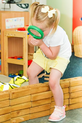 Bright young girl exploring a classroom with a magnifying glass, engaging in STEM play at Thrive Childcare, fostering curiosity and early learning skills.