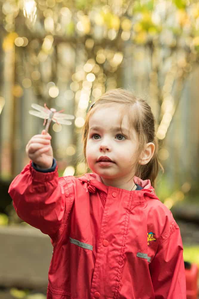 Bright young girl in colourful raincoat exploring outdoors, holding a dragonfly, representing happy, caring childcare environment at Thrive Childcare, nurturing early childhood development.