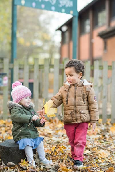 A young boy helping a little girl collect autumn leaves outside at Thrive Childcare, highlighting outdoor play and child development.