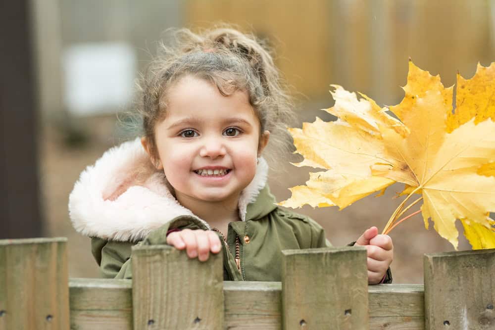 Bright young girl smiling outdoors with autumn leaves, promoting quality childcare and early childhood education at Thrive Childcare.