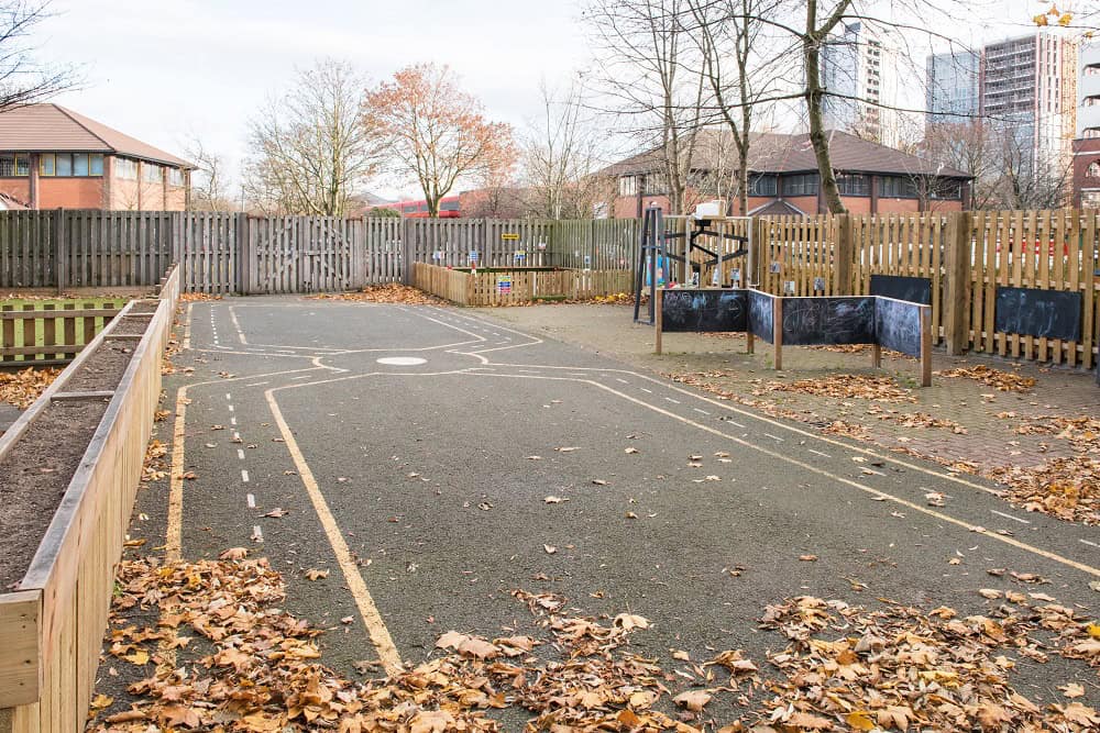 A safe outdoor play area at Thrive Childcare featuring a sports court with leaves scattered on the ground, surrounded by wooden fences and play equipment, ideal for children's outdoor activities.