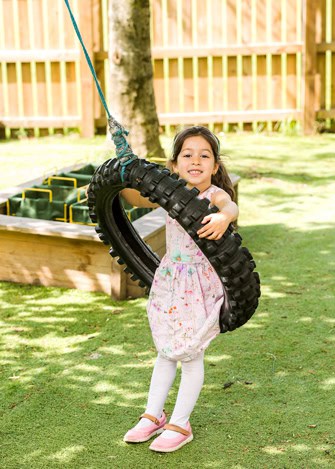 Bright smiling girl playing on a tire swing in a colourful childcare outdoor play area at Thrive Childcare centre in the UK, promoting happy childhood and outdoor activities for early years development.