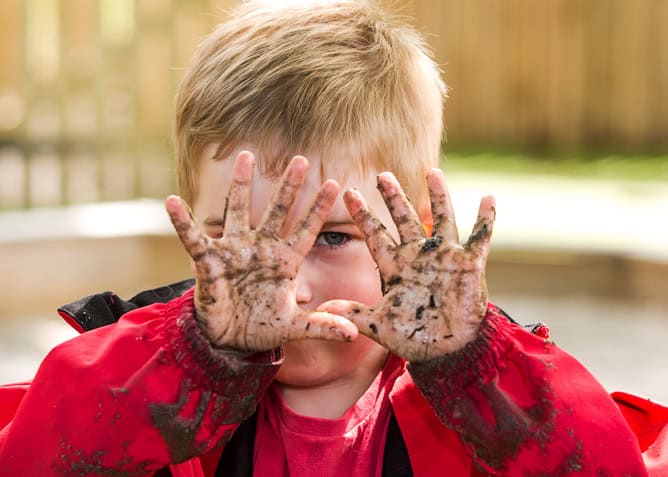 Colourful young child with muddy hands and face, enjoying outdoor play at Thrive Childcare. Engaging in creative, messy activities to promote preschool development and sensory exploration.