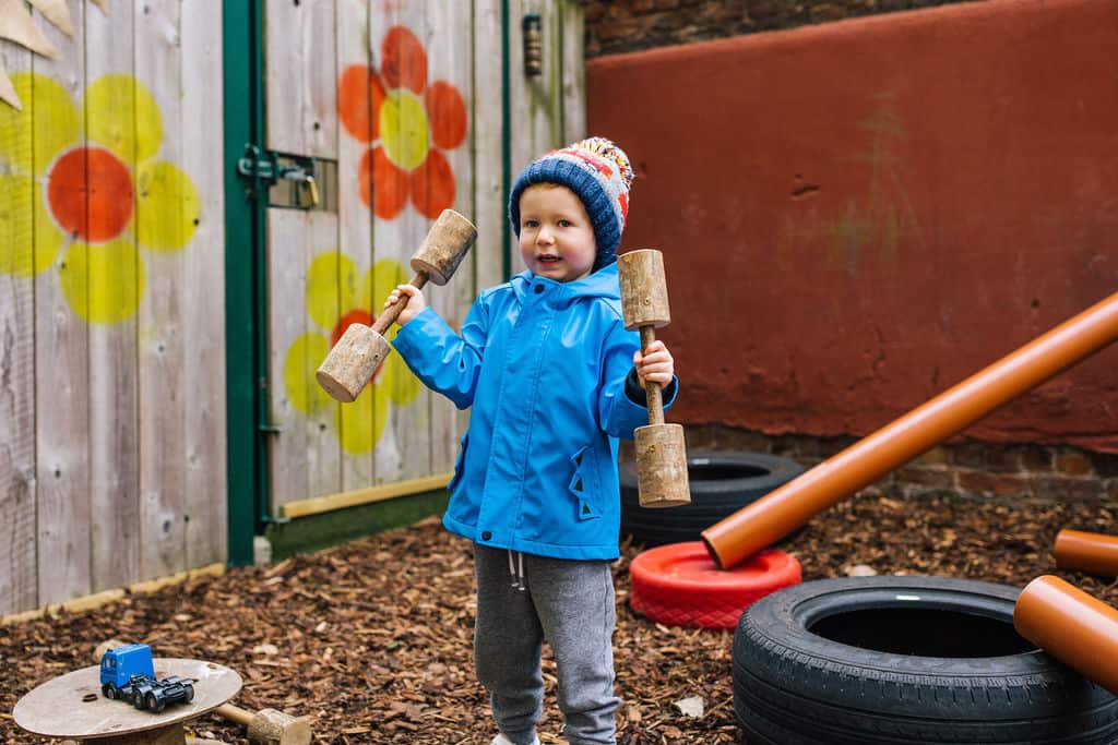 Junior child playing with a wooden hammer outdoors at Thrive Childcare, engaging in active, outdoor play activities in a safe, colourful playground environment designed for early childhood development.