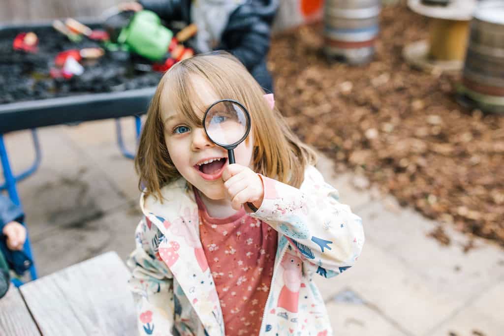 Optimised ALT text: Young girl with blue eyes holding a magnifying glass, exploring outdoors at Thrive Childcare, highlighting early childhood education and outdoor learning in a safe, nurturing environment.