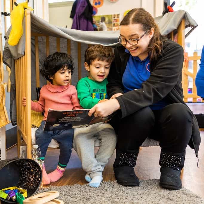 1. Happy childcare educator reading a book to young children in a colourful preschool setting, promoting early childhood learning and development.