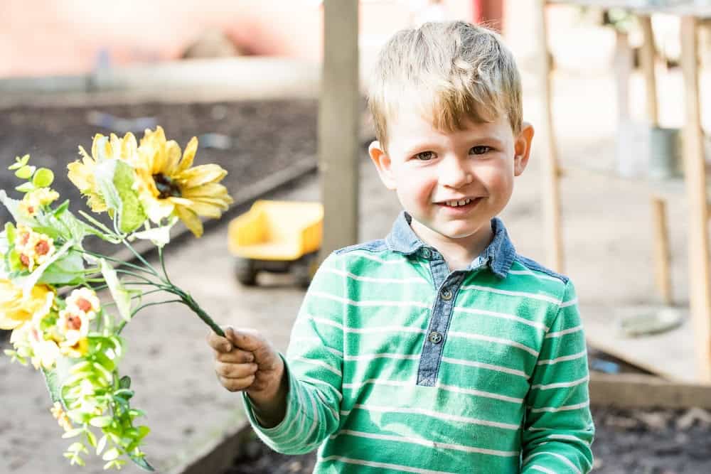 Bright young boy smiling outdoors at Thrive Childcare holding a colourful flower bouquet in a sunny playground setting.