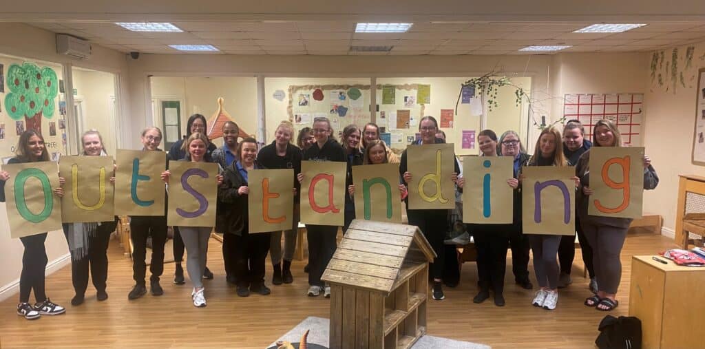 Bright group of childcare staff holding colourful "outstanding" signs at Thrive Childcare, showcasing commitment to high-quality early childhood education and child development in a nurturing environment.
