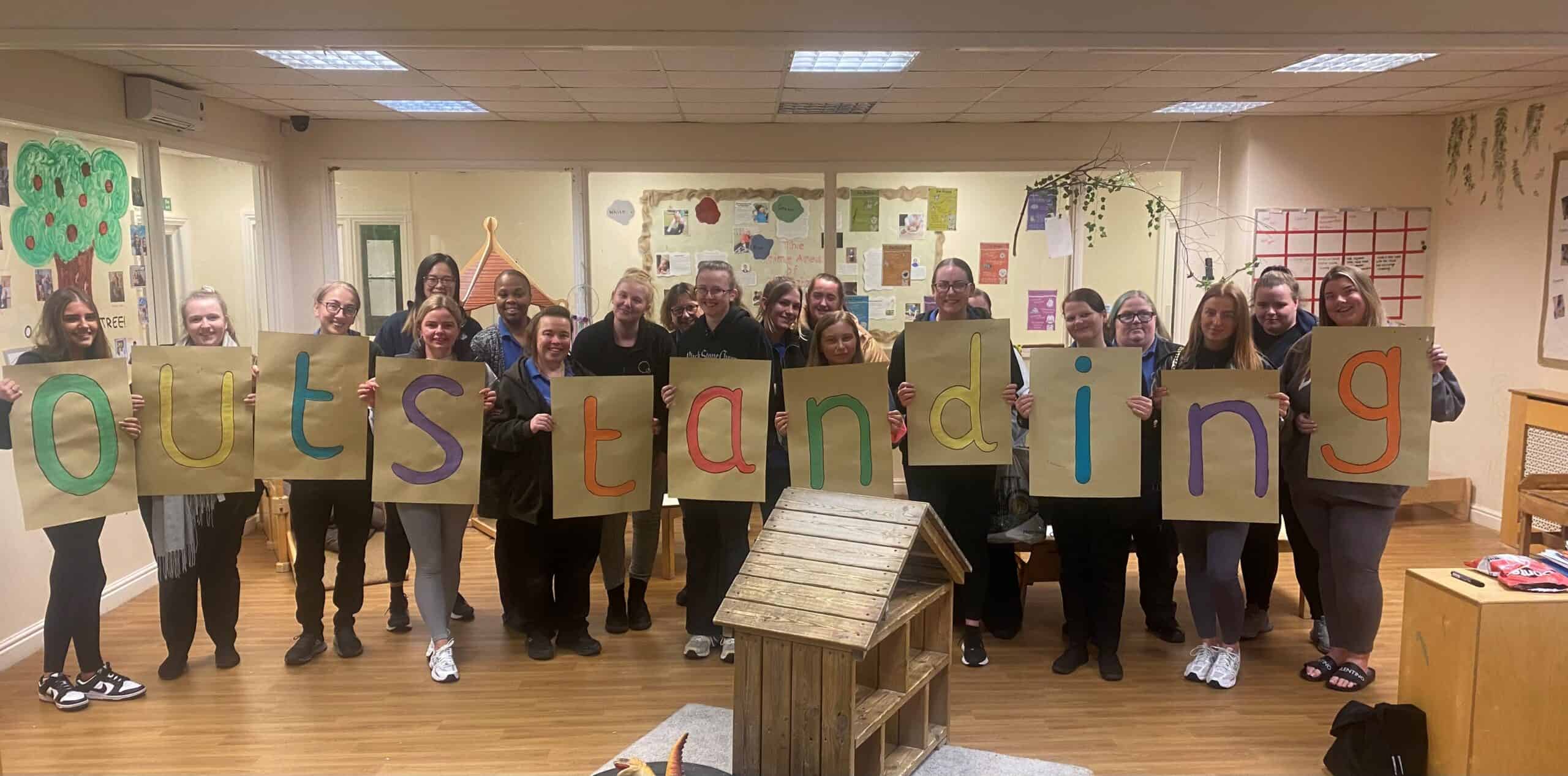 Bright group of childcare staff holding colourful "outstanding" signs at Thrive Childcare, showcasing commitment to high-quality early childhood education and child development in a nurturing environment.