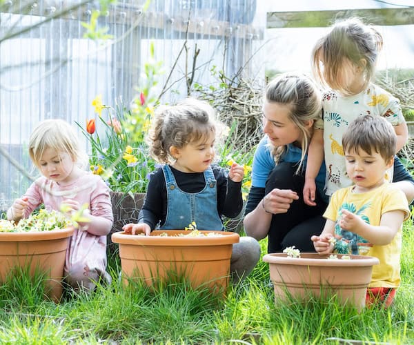 Bright young children planting flowers in a garden with caregiver, fostering outdoor learning and development at Thrive Childcare, a trusted early years education provider.