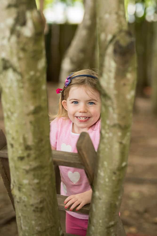 Vibrant young girl smiling behind a wooden play structure at Thrive Childcare, promoting happy childhood, outdoor play, and child development.