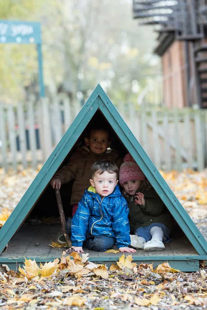 Children playing in a colourful outdoor wooden playhouse at Thrive Childcare, surrounded by autumn leaves, promoting early childhood development and outdoor learning activities.
