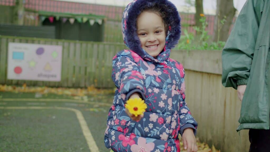 Bright smiling child in colourful jacket holding yellow flower at Thrive Childcare outdoor play area in England, promoting happy early childhood development and outdoor activities.