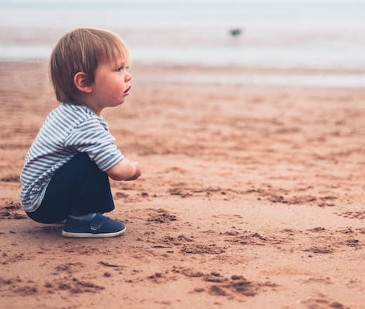 Young child sitting on sandy beach, gazing at the water, enjoying outdoor play and nature, representing early childhood development and outdoor activities at Thrive Childcare.
