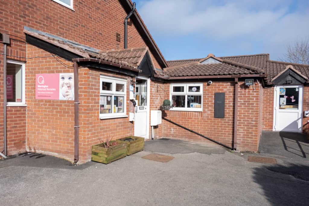 Bright red brick childcare centre façade with white-framed windows, entrance door, and TB signs, surrounded by clear blue sky, representing a safe and caring environment for young children’s early education and development.