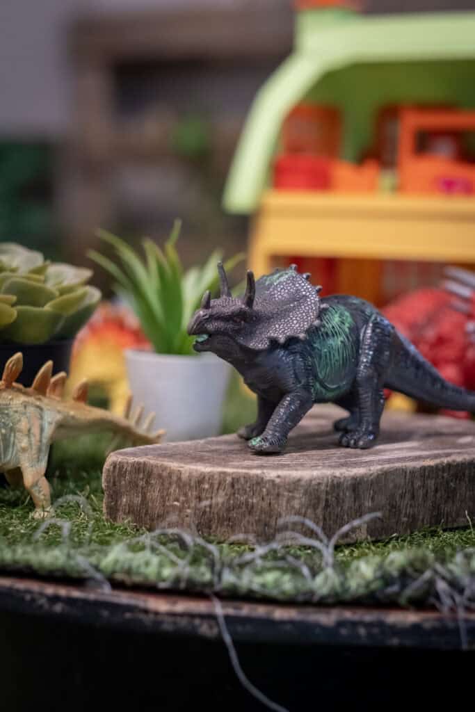 Colourful dinosaur toy on wooden log with plants and toys in the background at Thrive Childcare, promoting a fun and engaging environment for early childhood development and learning.