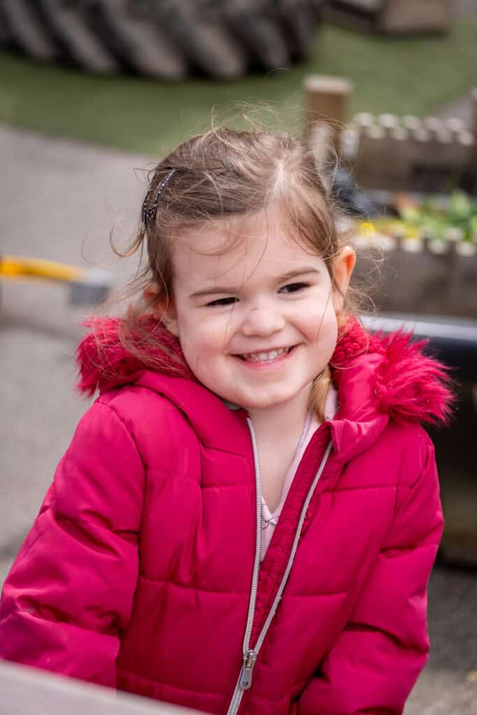 Smiling young girl in a bright pink winter coat at Thrive Childcare outdoor play area, showcasing child-friendly environment, safety, and engaging activities for early childhood development and care.