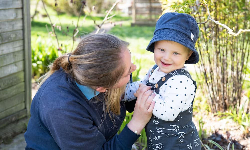 Bright cheerful young child in a blue sunhat and starry patterned dungarees enjoying outdoor play with a caring adult, showcasing nurturing childcare services at Thrive Childcare.