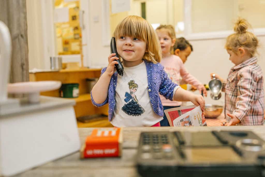 A young girl with red hair talking on a phone at Thrive Childcare, engaging in pretend play and learning activities with her friends in a colourful, nurturing environment.