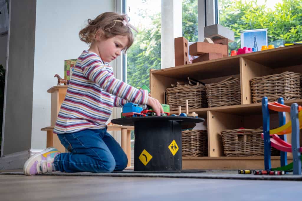 Young girl playing with toy trains on a black circular platform in a colourful childcare playroom with wooden shelves and natural light.