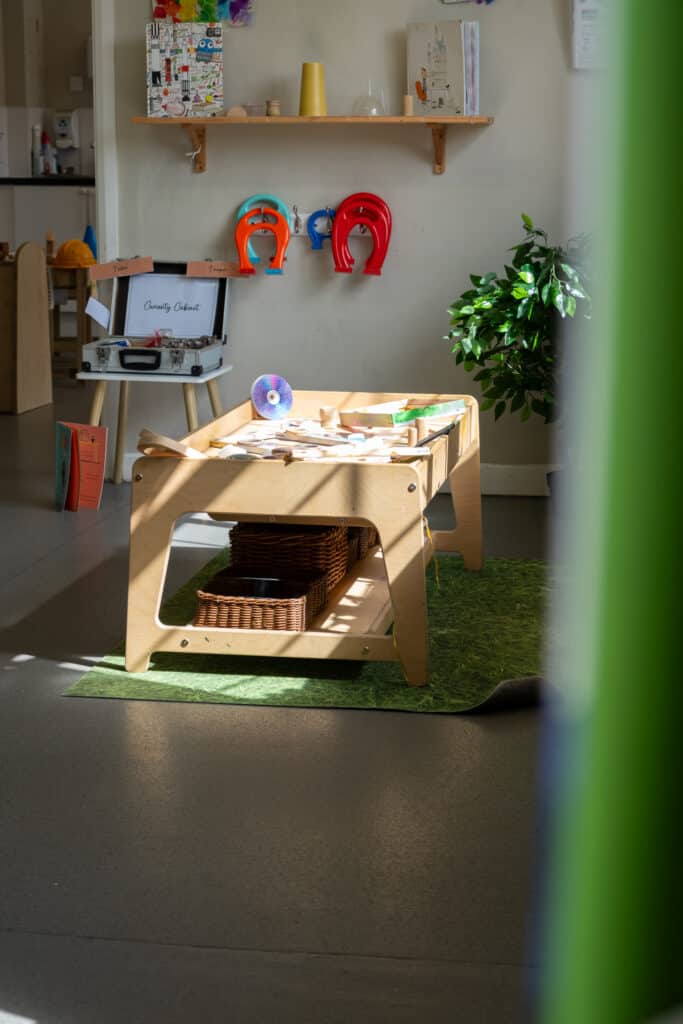 Cozy baby play area with wooden furniture, educational toys, colourful wall hooks, and natural sunlight at Thrive Childcare, fostering learning and development for young children.
