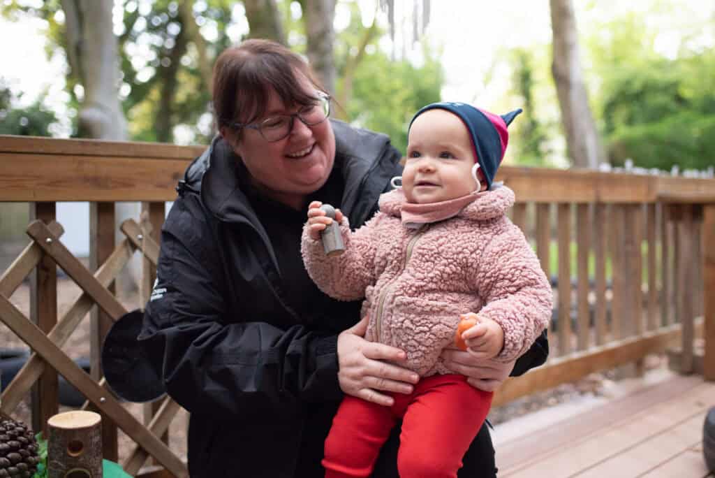 Bright cheerful child with a pink fleece jacket and colourful hat enjoying outdoor play at Thrive Childcare with a smiling caregiver.