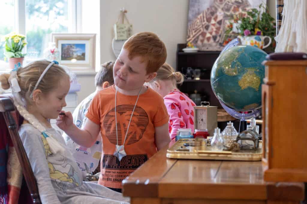 A young boy gently applying makeup to a girl with blonde hair in a colourful classroom setting, surrounded by educational toys, plants, and artwork, representing engaging childcare activities at Thrive Childcare.