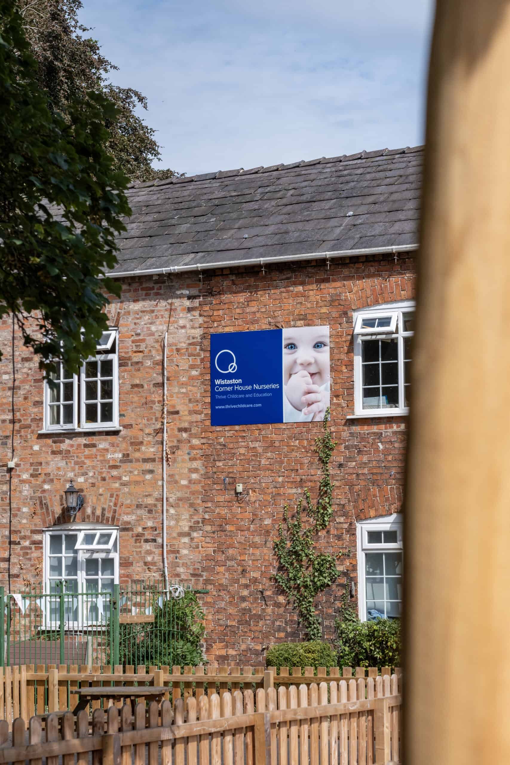 Bright brick nursery building exterior with Thrive Childcare signage, featuring a child's happy face, outdoor garden, and fencing, representing quality childcare and early education services in Wistaston.