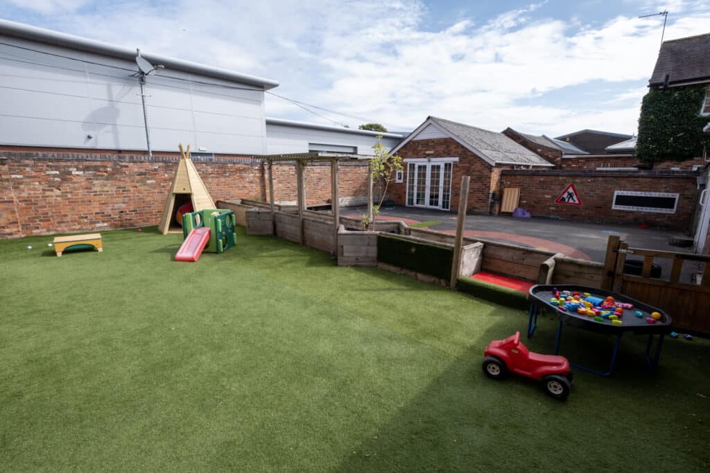 Bright outdoor play area at Thrive Childcare with artificial grass, children’s toys, climbing frame, slide, sandpit, and outdoor learning space, fostering early childhood development and safe play.
