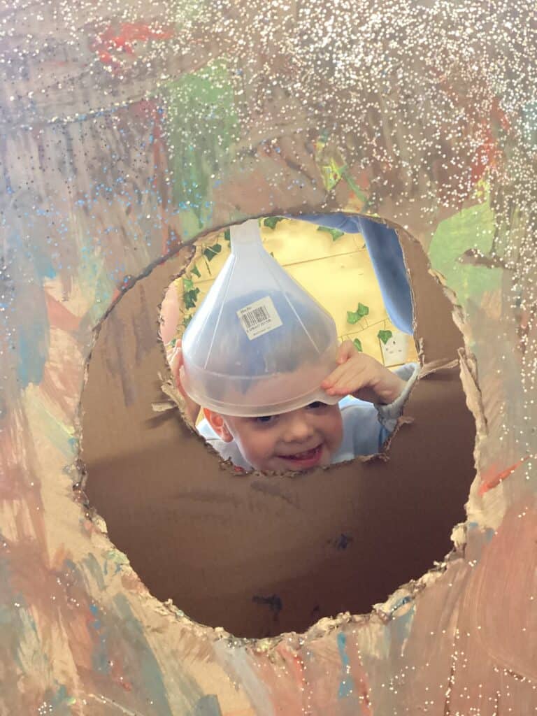 Bright smiling child peeks through a cardboard cutout hole, wearing a clear plastic cone, at Thrive Childcare, highlighting safe, engaging activities for early childhood development in a nurturing environment.