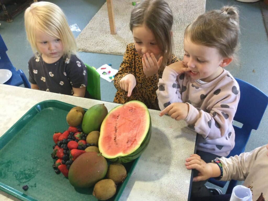 Bright preschool children examining a variety of fresh fruits including watermelon, strawberries, blueberries, and kiwis at Thrive Childcare, promoting healthy eating and sensory play for early childhood development.