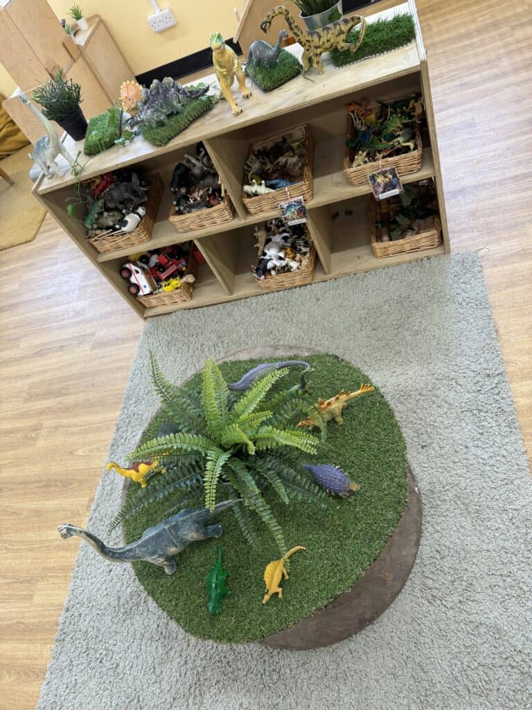Colourful dinosaur toys and plants on a wooden shelf, with a round grass area containing more dinosaur figures, creating an engaging educational play space for young children at Thrive Childcare.
