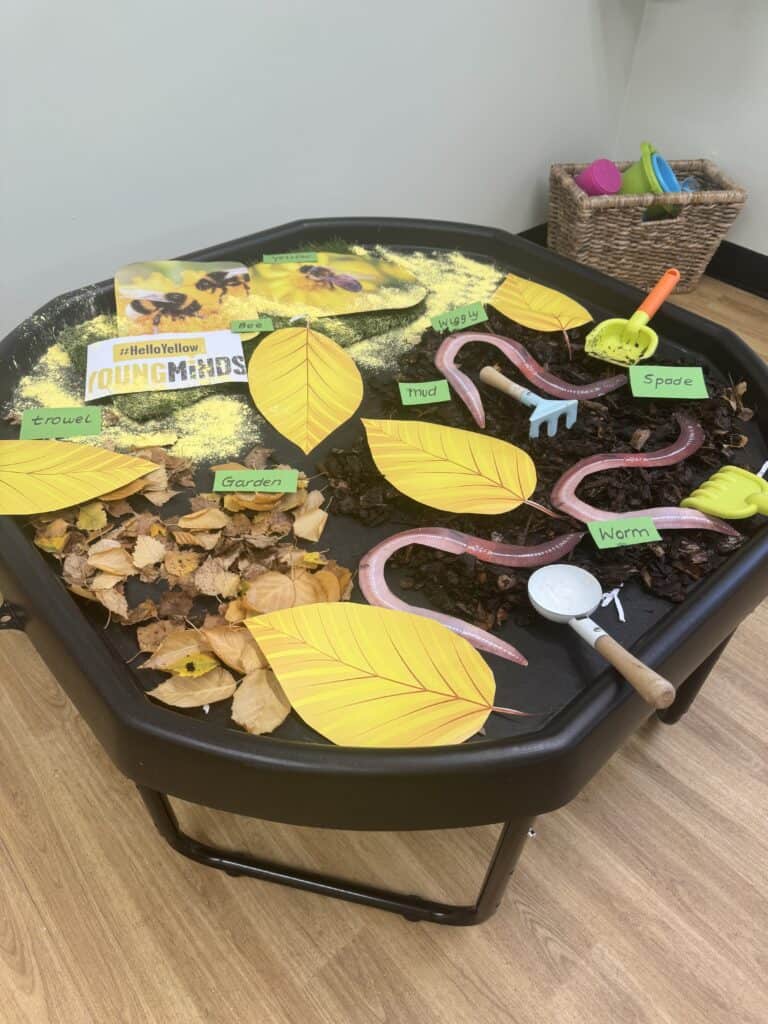Bright educational play table with autumn-themed sensory activity, including leaves, soil, plastic worms, and labelled tools, suitable for toddler early learning in a childcare setting.