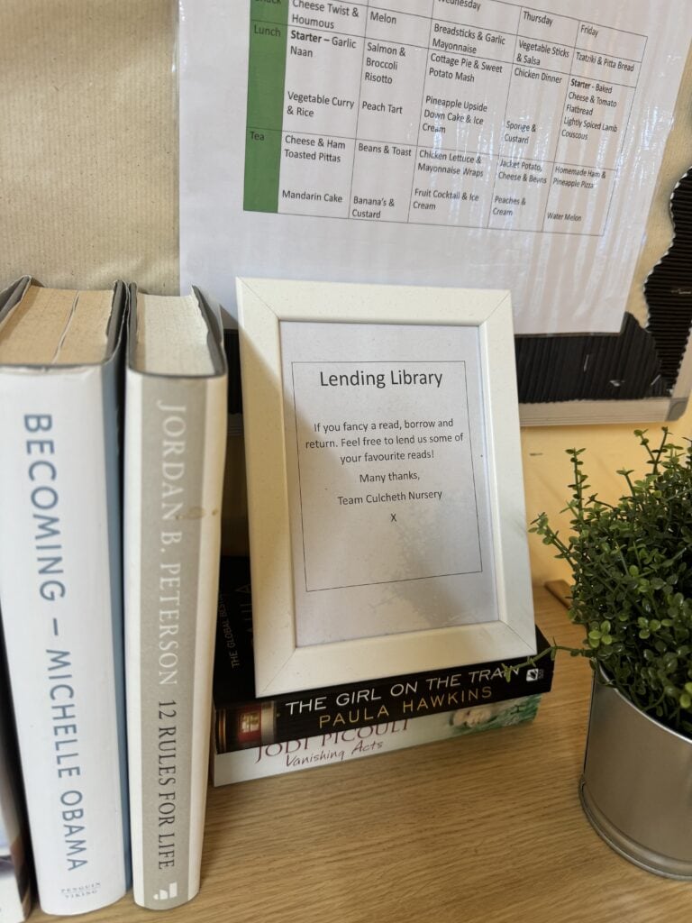1. Shelves with books and a framed sign for the Lending Library at Thrive Childcare, encouraging children to borrow and return books by team Culcheth Nursery.