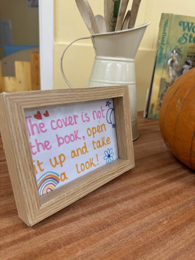 Bright framed sign with a colourful message about opening a storybook, surrounded by a pumpkin, a book, and a cream jug on a wooden table in a cheerful childcare environment.