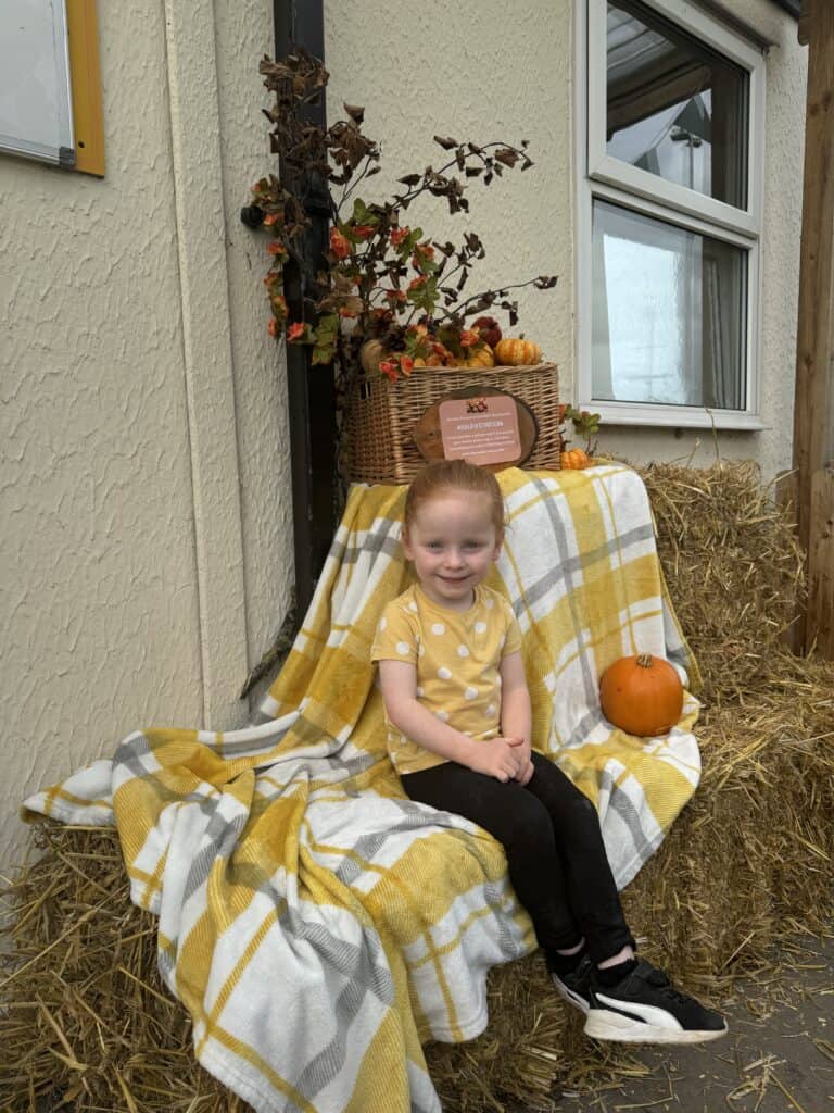 Freshly dressed young girl sitting outdoors on haystack with pumpkins, fall decor at Thrive Childcare, promoting a safe and nurturing environment for children's development.