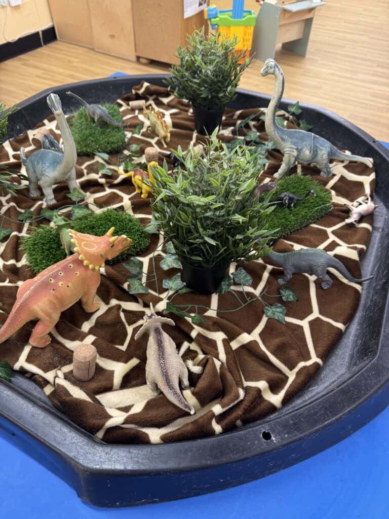 Colorful dinosaur toys in a classroom sensory play area with plants and soft fabric, promoting imaginative learning and environmental exploration for early childhood development.