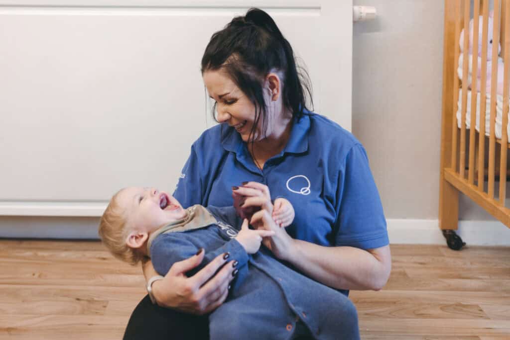 Joyful childcare worker playing with smiling toddler in nursery, creating a happy learning environment, professional childcare, early childhood development, Thrive Childcare services, engaging activities for young children.