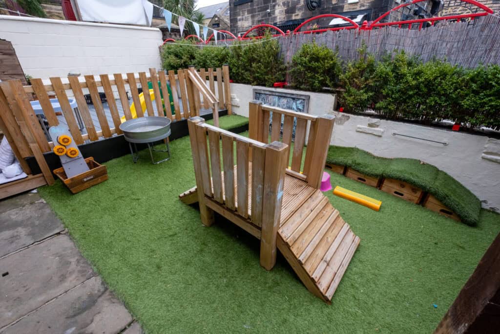 Bright outdoor toddler play area at Thrive Childcare with wooden ramps, sensory toys, and artificial grass, providing a safe and engaging environment for early childhood development.