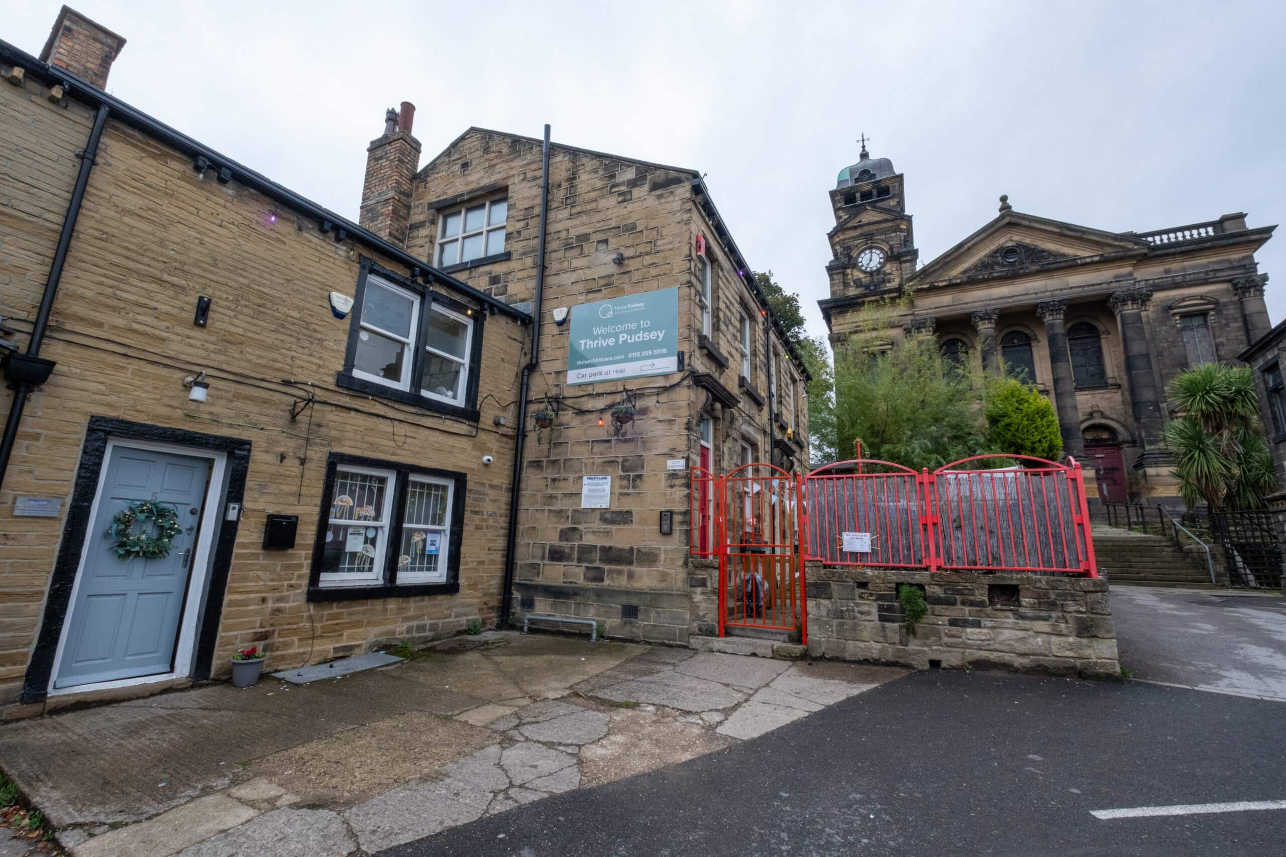 Bright yellow brick building housing Thrive Childcare centre, with welcoming entrance, situated next to historic church on a quiet street in Pudsey, Leeds, offering quality early years education and childcare services.