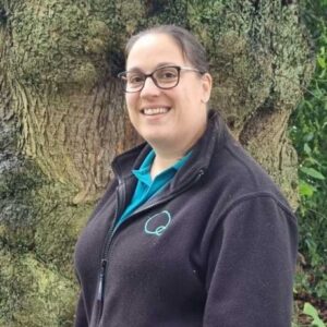 A smiling childcare provider outdoors, wearing a Thrive Childcare branded jacket near a large tree, promoting safe and nurturing early childhood education services.