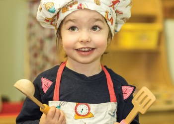 Bright cheerful young girl wearing a chef's apron and hat, smiling with cooking utensils in hands, in a colourful childcare environment, promoting early childhood education and fun activities at Thrive Childcare.
