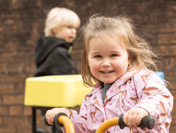 Bright smiling young girl riding a yellow tricycle at Thrive Childcare, showcasing happy children enjoying outdoor activities and play-based learning.