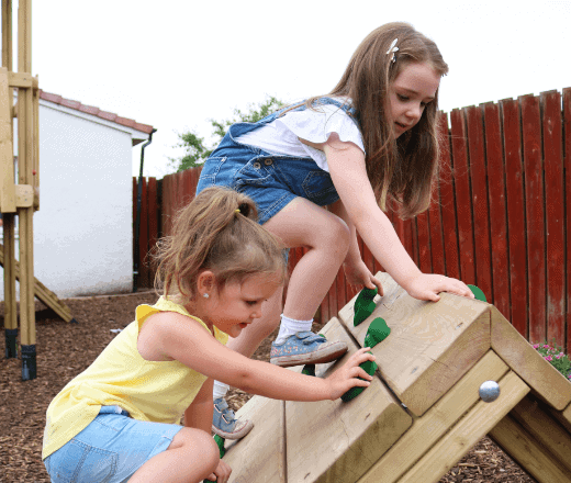 A smiling young girl and a younger girl playing together on a wooden outdoor climbing frame at Thrive Childcare.
