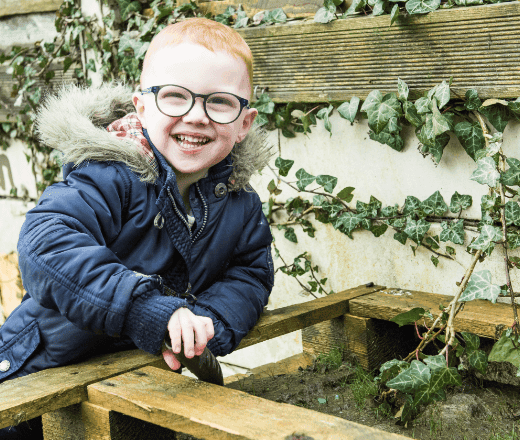 A happy young boy gardening outdoors, smiling, wearing glasses and a navy jacket, surrounded by green ivy, in a vibrant green outdoor setting.