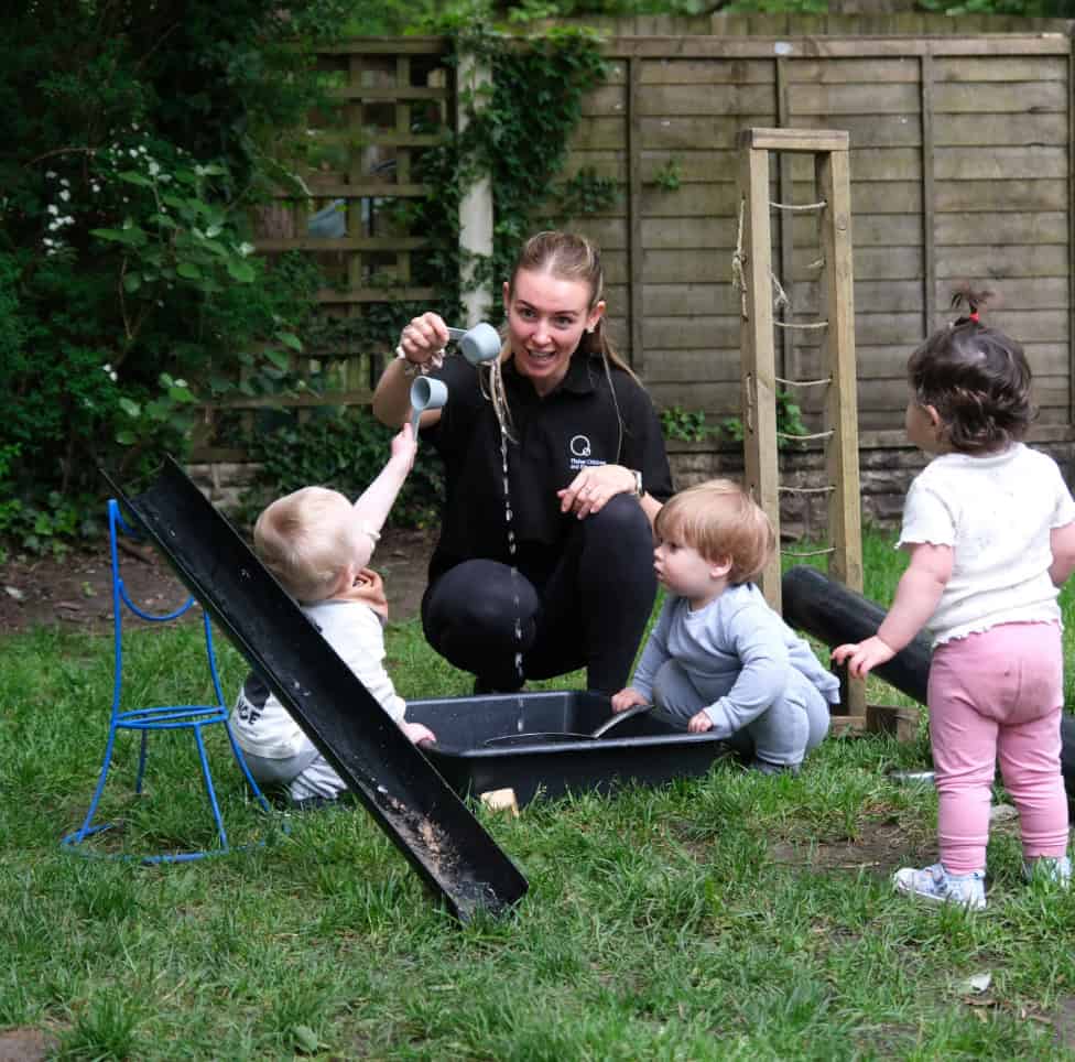 Young children playing outdoors at Thrive Childcare, supervised by a caregiver, engaging in water play and sensory activities in a safe, natural garden environment.