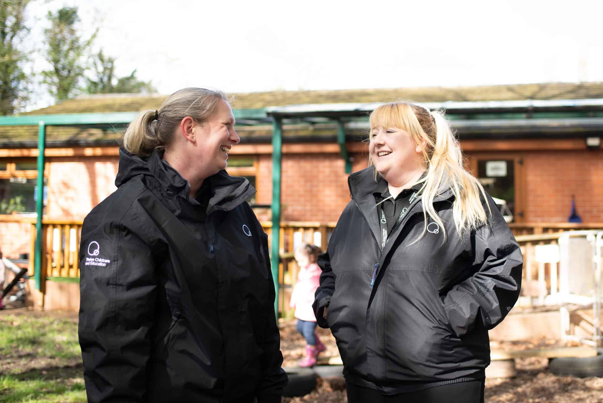 1. Two joyful childcare staff members in black Thrive Childcare jackets engaging outdoors with children in a sunny garden setting.