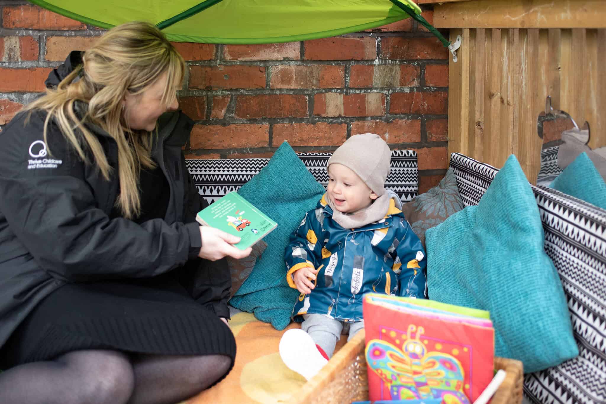 1. Friendly childcare professional reading a colourful picture book to a happy toddler in a cosy outdoor play area at Thrive Childcare.