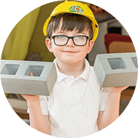 Bright young boy in safety helmet holding two concrete blocks, demonstrating creativity and developing fine motor skills at Thrive Childcare, fostering early learning and fun.