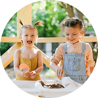 A joyful young girl and boy playing outdoors on a wooden deck, engaging in creative activities with soil and tools at Thrive Childcare, promoting early childhood learning and development.